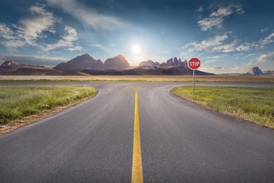 Crossing with stop sign towards the sun and mountain peaks with yellow line on asphalt. Success and vision concept.