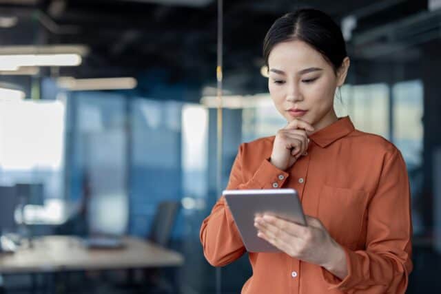 Serious Asian businesswoman pondering over digital tablet in office