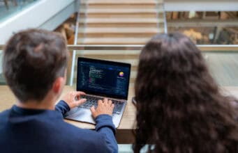 Two software developers working together on coding and system management on a laptop. Professional programmers collaborating on web development and software engineering in a modern tech office workspace.
