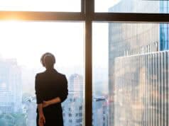 Faculty Perspective: The Leadership Transition No One Prepares You For Professional standing by a window overlooking a city skyline.