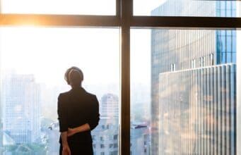 Faculty Perspective: The Leadership Transition No One Prepares You For Professional standing by a window overlooking a city skyline.
