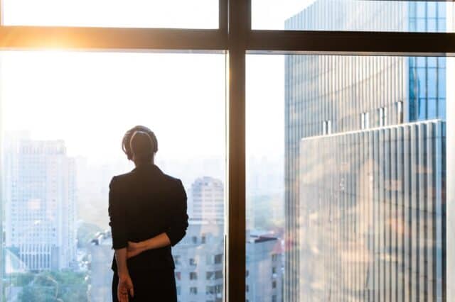 Thoughtful Leadership Stock Image-2274395065-resized Professional standing by a window overlooking a city skyline.
