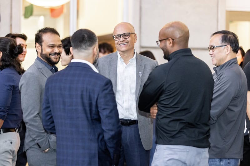 Goizueta Dean Saby Mitra, wearing a gray blazer and glasses, smiles while speaking with a group of attendees at an indoor anniversary celebration.