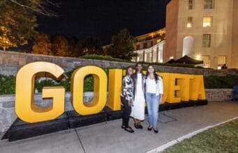 Goizueta Celebrates 2026 Homecoming and Reunion Weekend Three women pose in front of large gold GOIZUETA letters at an outdoor evening celebration on Emory’s campus.