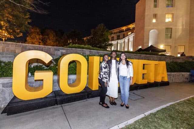 Homecoming 2026_MSBA 10 YR_7I6A0442 - resized Three women pose in front of large gold GOIZUETA letters at an outdoor evening celebration on Emory’s campus.